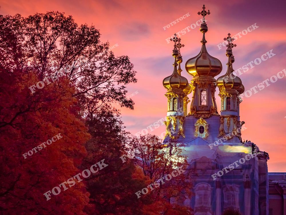 Фотография показывает золотые купола Екатерининского дворца в Царском Селе на фоне закатного неба. Вечерний свет подчеркивает величие архитектуры и насыщенные цвета осенней листвы вокруг. Екатерининский дворец, известный своей роскошной отделкой и архитектурой в стиле барокко, является одной из главных исторических достопримечательностей Санкт-Петербурга. Закатные оттенки и золото куполов создают атмосферу спокойствия и величия.
